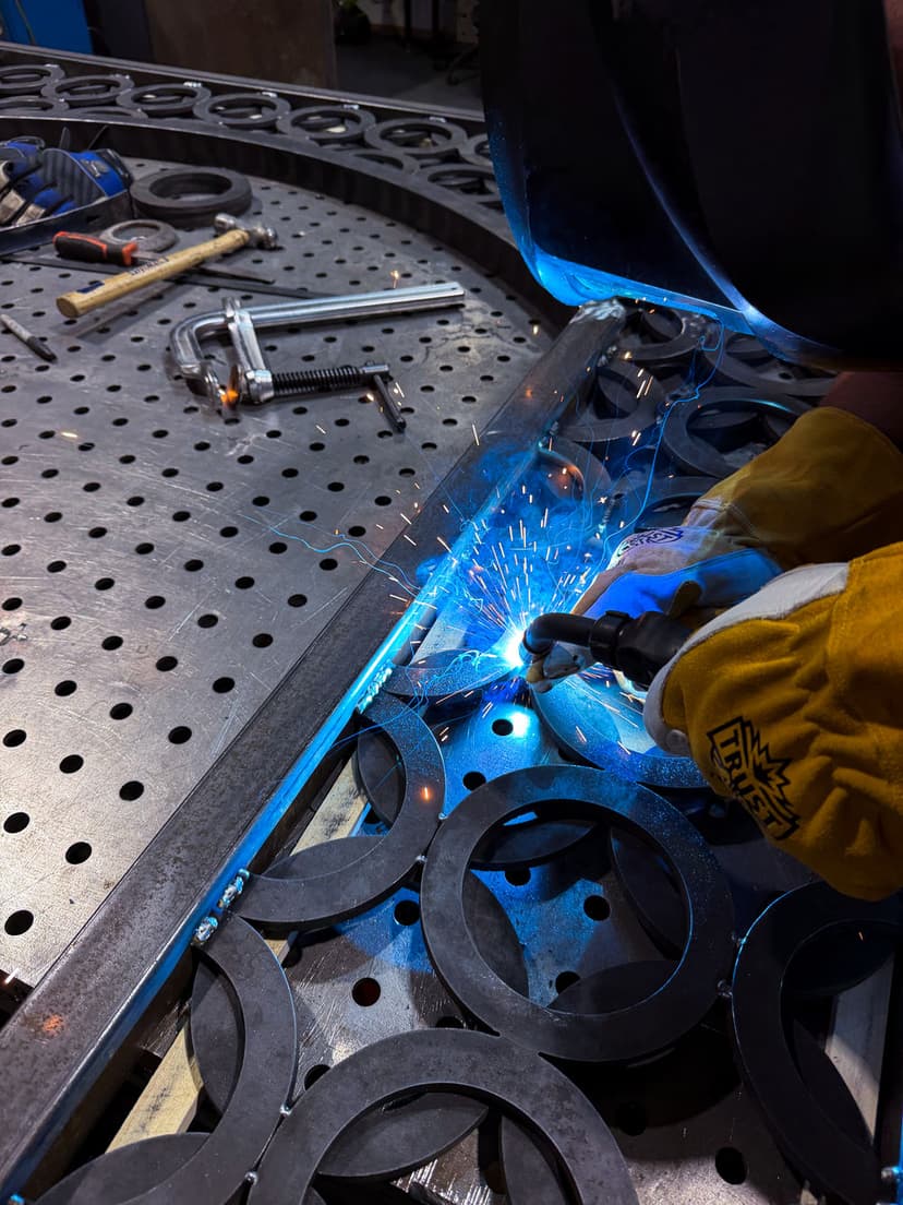 Welder using a TIG torch on metal, sparks flying, with tools and fixtures in the background.