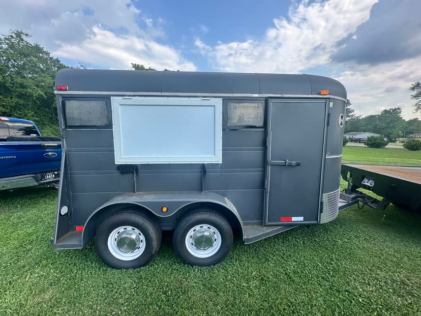 Black horse trailer with dual wheels, parked on grass with blue sky and clouds in the background.