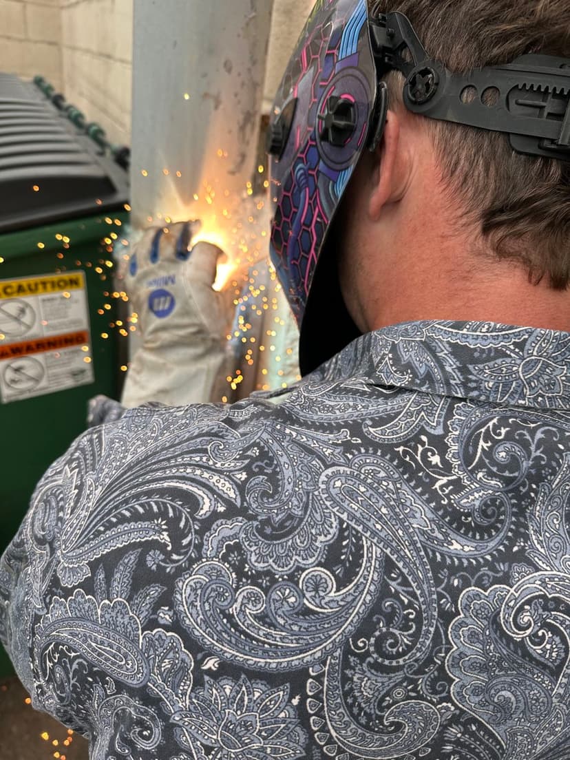 Person wearing a helmet and gloves welding near a green dumpster, sparks flying.